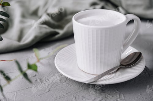 Preview: White cup of frothy milk coffee on a textured table with a spoon and green leaves