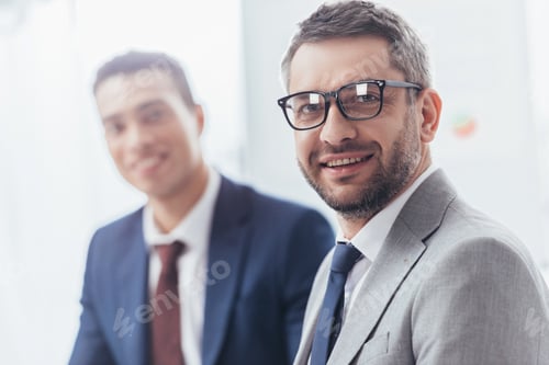 Preview: handsome bearded businessman in eyeglasses smiling at camera, young colleague standing behind
