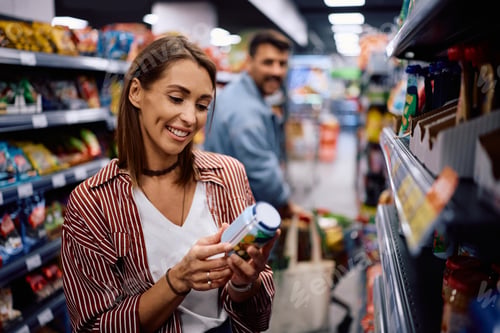 Preview: Happy woman reading label on food jar while buying in supermarket.