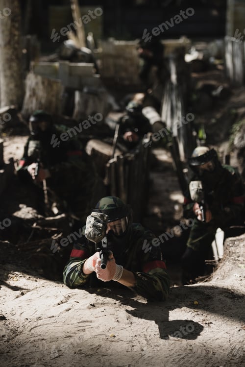 Preview: paintball team in camouflage and protective masks aiming by marker guns from ditch outdoors