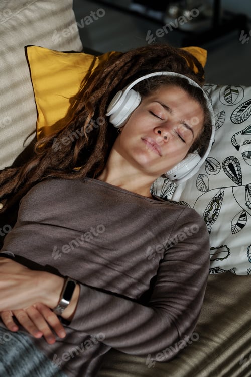 Preview: Pretty young relaxed woman with headphones lying on soft couch in dark room