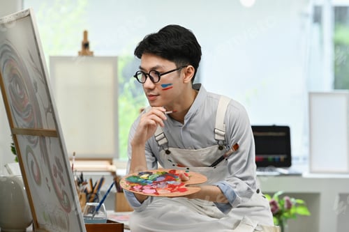 Preview: Pleased male artist holding palette and looking at his artwork with admiration.