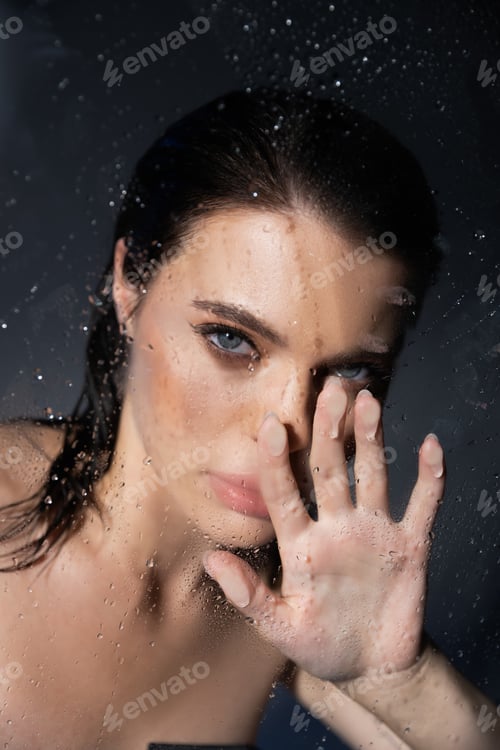 Preview: Young brunette woman with makeup touching wet glass on grey background