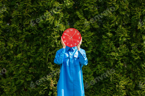 Preview: Stylish woman in blue shirt hold red clock on green trees background