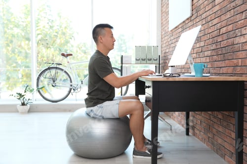 Preview: Young man sitting on fitness ball and using computer in office. Workplace fitness