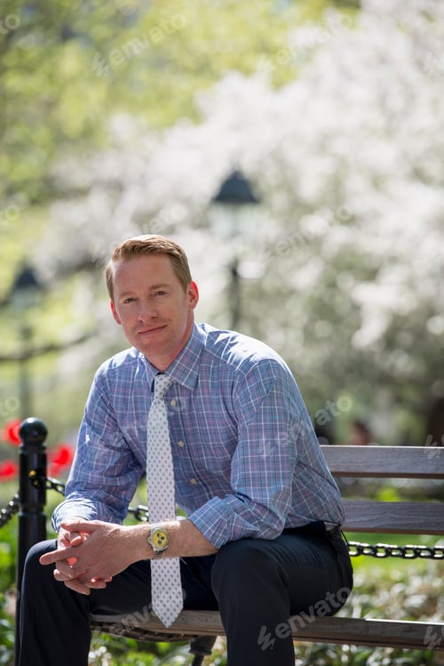 Preview: A businessman in a shirt with white tie, sitting on a park bench.