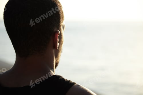 Preview: Back shot of handsome dark-skinned young athlete enjoying sunny summer days at ocean, contemplating