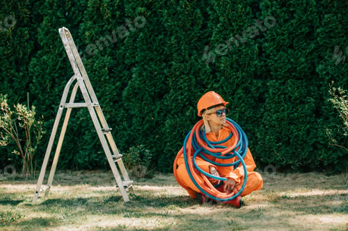 Preview: Stylish woman in orange suit, hardhat, pipes and sunglasses next to stepladder