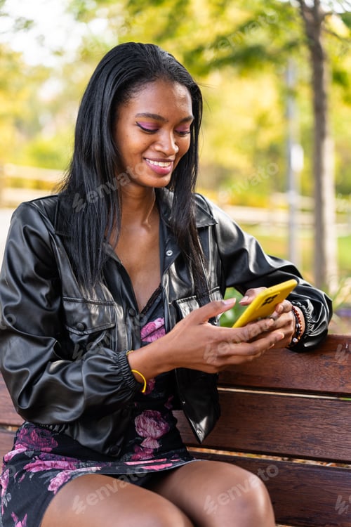 Preview: Modern black woman sitting in the park smiling with her mobile phone in her hand