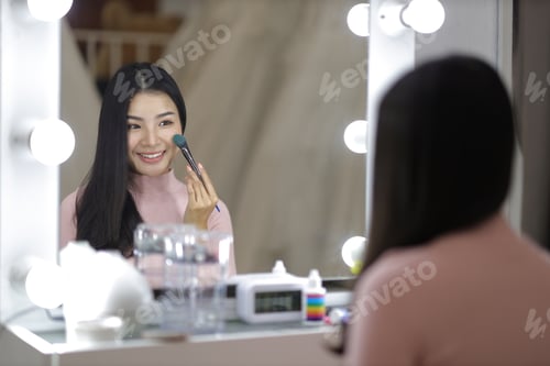 Preview: Cute young Asian woman looking at herself in the mirror while applying makeup.