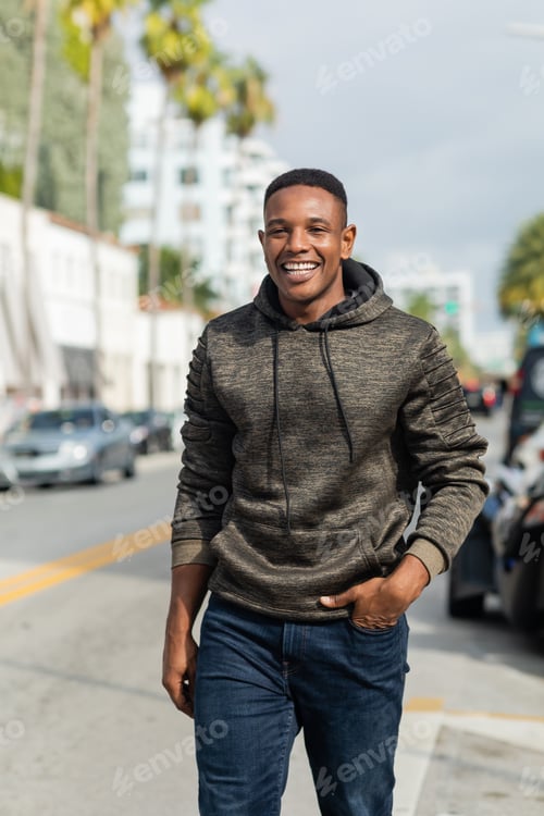 Preview: happy african american man in hoodie and jeans walking with hand in pocket on street in Miami