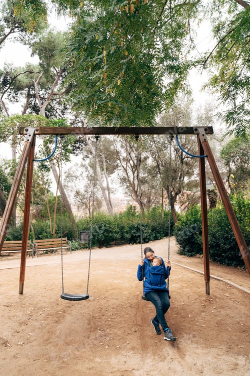 Preview: Mom in a blue warm sweater and jeans swinging on a swing with a tiny baby on her chest in the park