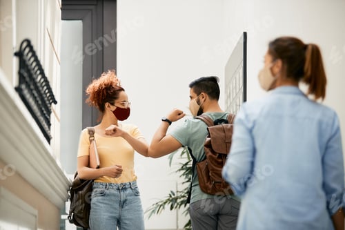 Preview: Happy college friends with face masks greeting with elbows in a hallway.