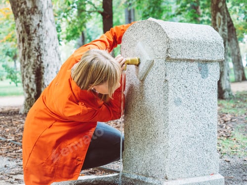 Preview: Caucasian girl with red gabardine drinks water from a fountain in a park in Madrid