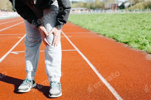 Preview: Woman runner suffering from knee pain holding her leg in stadium outdoors, close-up.