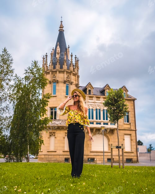 Preview: Portrait of a blonde woman in a yellow vintage outfit with a hat walking next to a castle