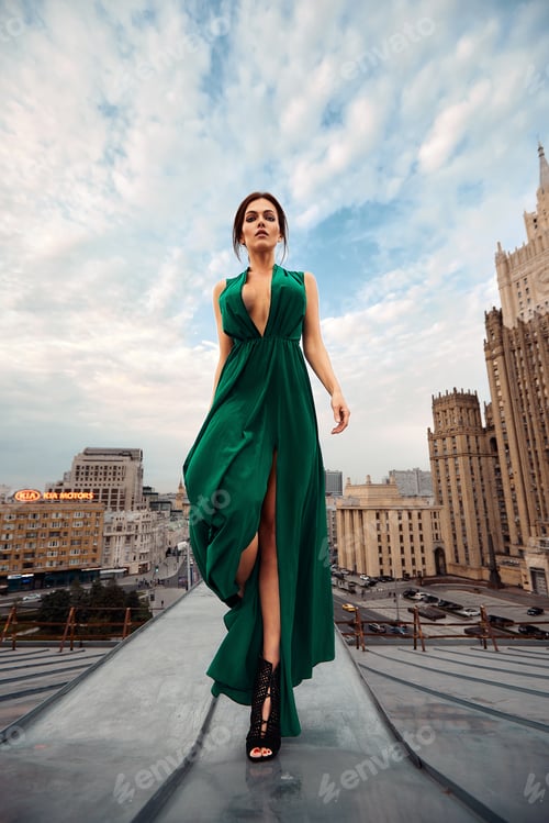 Preview: Attractive brunette girl in a long emerald dress posing on the catwalk on the roof against the