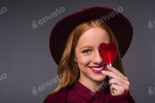 Preview: beautiful redhead stylish girl in hat with heart shaped lollipop, isolated on grey
