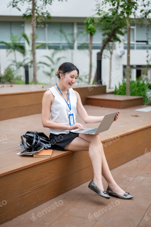 Preview: Asian office worker woman holding looking at laptop as sitting cross legged on wooden stairs in park