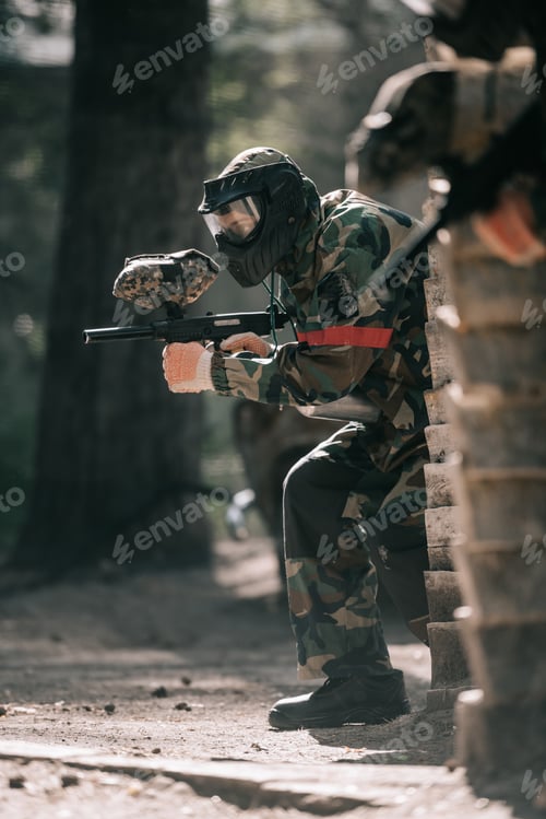 Preview: selective focus of male paintball player in goggle mask and camouflage aiming by paintball gun