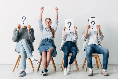 Preview: Smiling woman showing yes gesture near employees holding cards with question marks in office