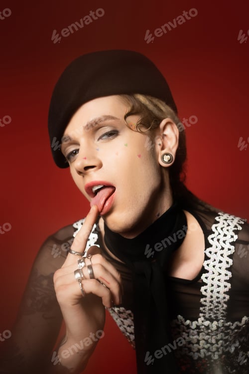 Preview: queer person in elegant attire sticking out tongue and looking at camera on red background