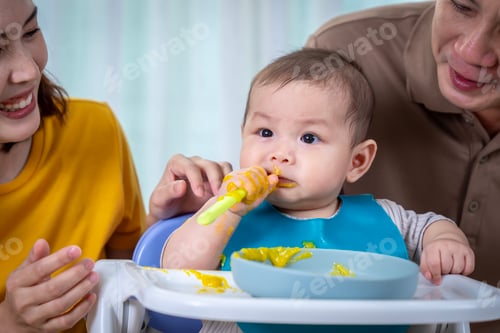 Preview: A baby is sitting in a high chair with a yellow mess on his face. The baby is wearing a green bib an