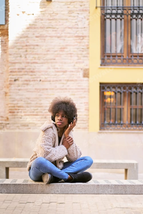 Preview: Confident ethnic teen sitting with crossed leg on stone bench while listening to music
