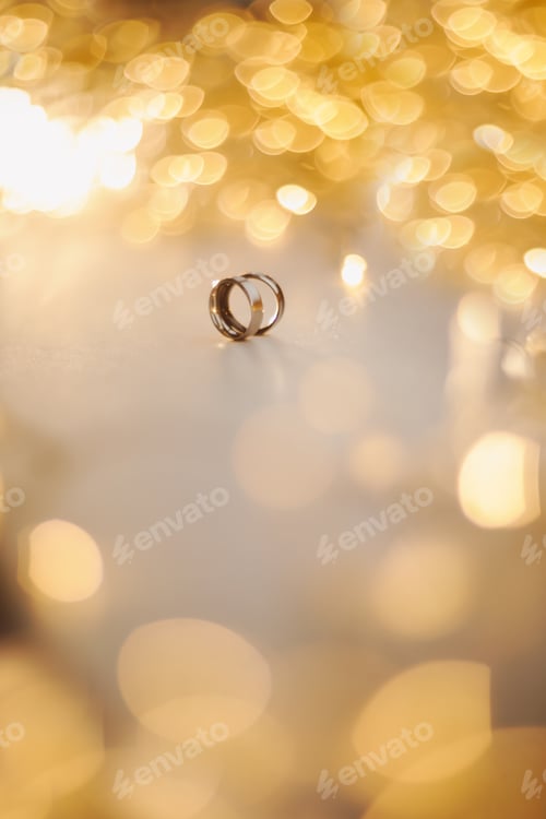 Preview: Wedding rings on blured bokeh glowing golden background. Symbol of love and romance on a textured