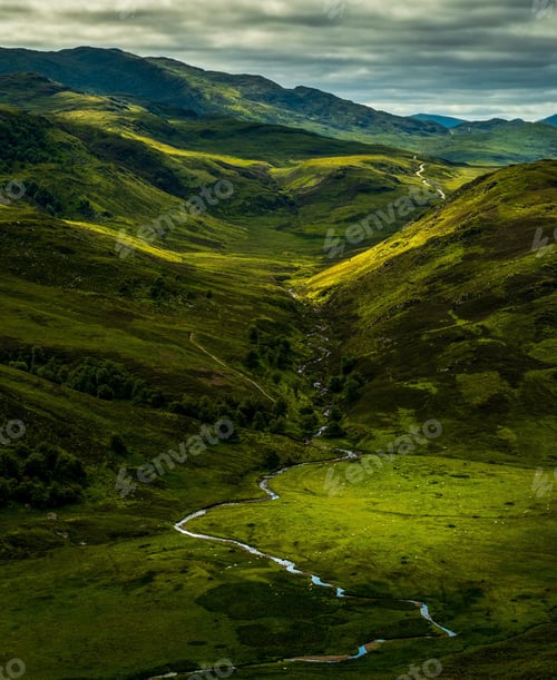 Preview: Small river flowing through green hills under a cloudy sky