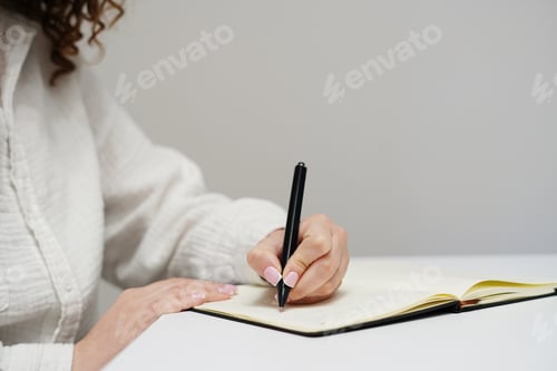 Preview: A woman in a white shirt is writing in a notebook with a black pen on a clean white desk