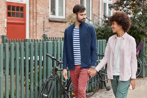 Preview: Photo of lovely interracial couple have outdoor walk, keep hands together and look at each other wit