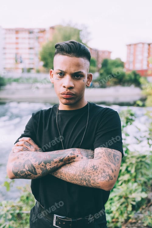 Preview: young hispanic tattoed man outdoor in the city, resolute with arms folded