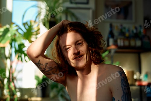 Preview: Shirtless young man with tattoos smiles warmly in sunlit room.