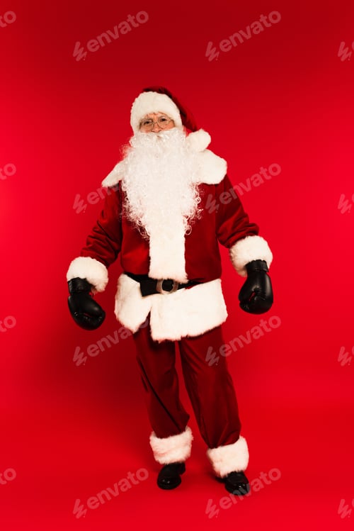 Preview: Full length of santa claus in costume and boxing gloves standing on red background