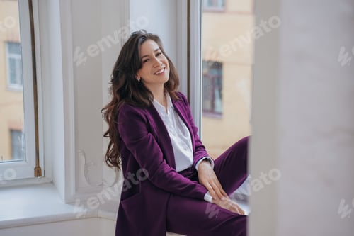 Preview: Gorgeous brunette hispanic woman in violet suit sits on windowsill smiles looks at camera, satisfied