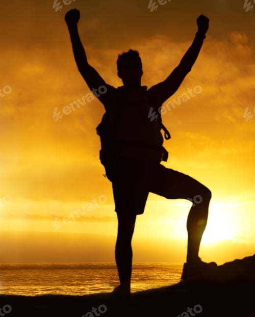 Preview: Conquering the mountain. Silhouette of a hiker standing on a mountain with his arms raised.