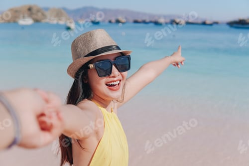 Preview: Cheerful young Asian woman holding hands on Pink Beach, Labuan Bajo while pointing to the scenery.