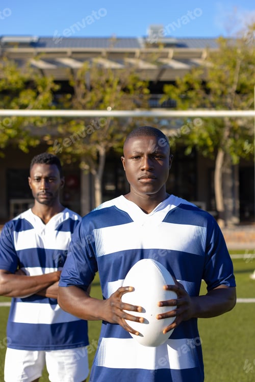 Preview: Two African American young male athletes, holding a rugby ball, standing on field outdoors