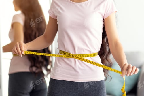 Preview: Unrecognizable Indian lady measuring her waist with tape in front of mirror at home, closeup