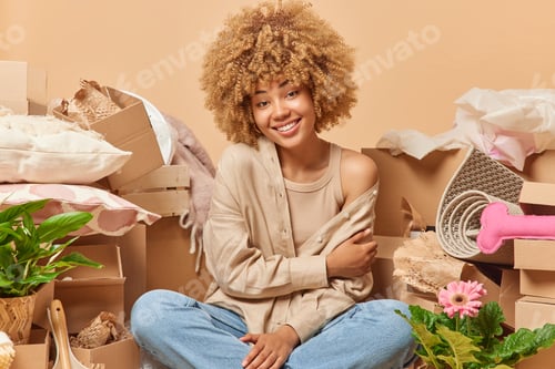 Preview: Positive curly haired woman sits crossed legs wears beige shirt and jeans poses around carton boxes