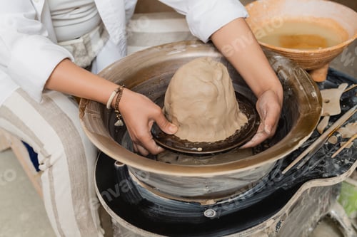Preview: Cropped view of young female artisan in workwear working with clay on pottery wheel, tools and bowl