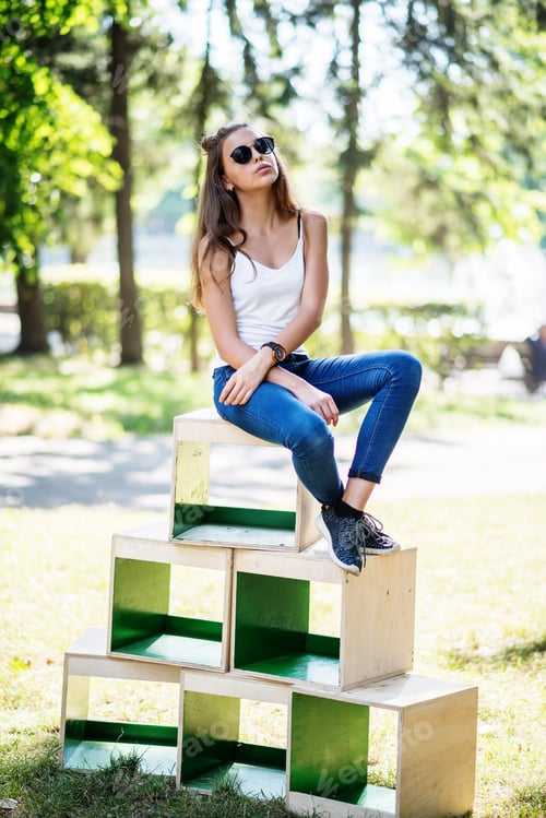 Preview: Portrait of a fashionably dressed girl posing on wooden boxes in the park on a sunny day