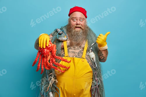 Preview: Excited bearded seaman points away on blank space wears yellow gloves and overalls holds big red oct