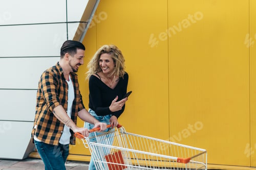 Preview: Couple using smartphone and holding shopping cart while outside of the supermarket or mall