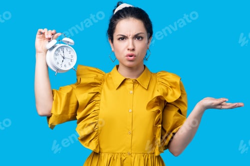 Preview: Woman in a mustard dress holding a clock and looking uncertain