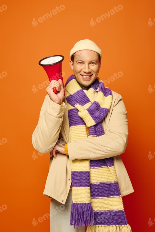 Preview: Cheerful young man with tattoos holding a megaphone in vibrant orange setting