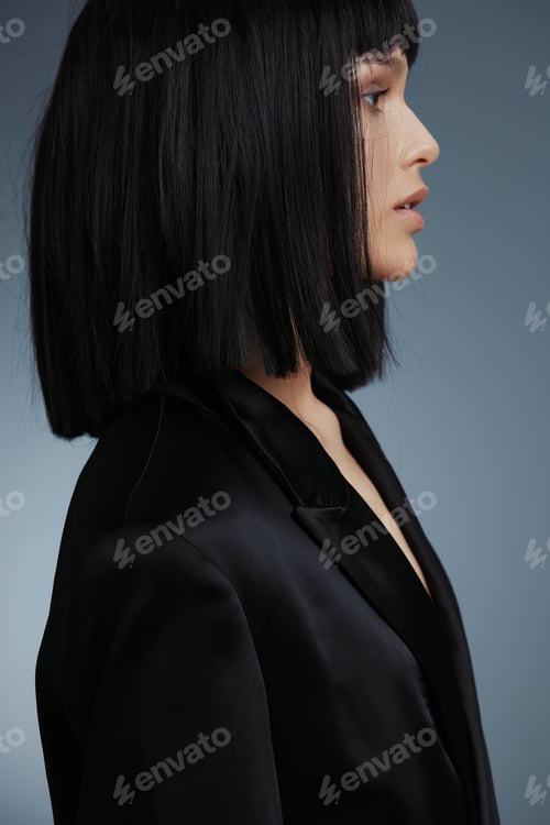 Preview: Blackhaired woman in a black suit looking sideways in a studio shot, set against a gray background