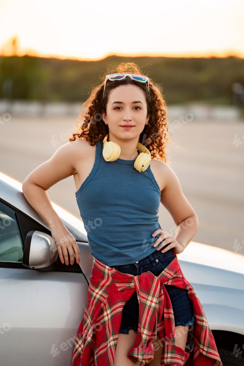 Preview: Curly-haired woman leaning on car with headphones, sunset backdrop.