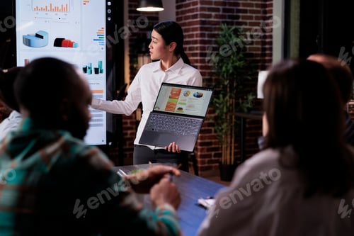 Preview: Asian woman presenting sales strategy to coworkers working overtime at the office